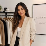Woman in beige blazer and white top posing in fashion boutique with clothing rack and Camila Ro signage behind her