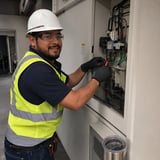 Electrician in white hard hat and yellow safety vest working on an electrical panel with black gloves