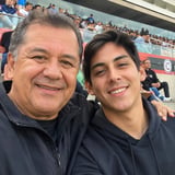 Two men smiling at camera at a crowded baseball stadium, wearing dark jackets with spectators visible in background