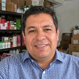 Man wearing blue checkered shirt smiling at camera in front of shelves with products and boxes