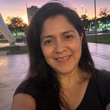 Woman smiling at camera in front of a public plaza during sunset with buildings and trees visible in the background