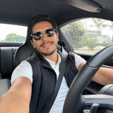 Man with dark hair and sunglasses sitting in a convertible car, wearing a white shirt and black vest, holding the steering wheel